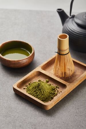 selective focus of green matcha powder and bamboo whisk on wooden board near black teapot and bowl...