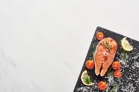 top view of raw salmon with seasoning and tomatoes on stone board on marble tableの写真素材