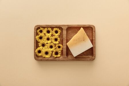 top view of brown wooden soapdish with flowers and piece of soap on beige background