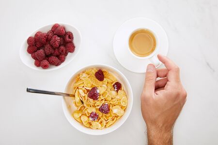 cropped image of man holding cup of coffee near bowl with cornflakes and plate with fresh raspberry...
