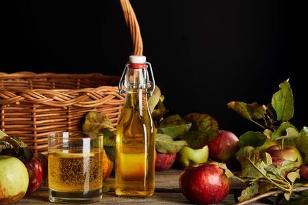 bottle and glass of homemade cider near wicker basket and apples isolated on black