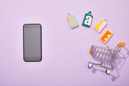 top view of smartphone near shopping bags with sale lettering falling into toy cart on violet
