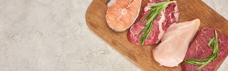 Panoramic shot of raw meat, fish and poultry with rosemary twigs on wooden cutting board on marble surface with copy spaceの写真素材