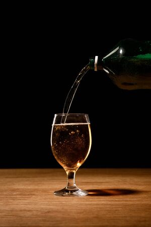 beer pouring from bottle in glass on wooden table isolated on blackの写真素材