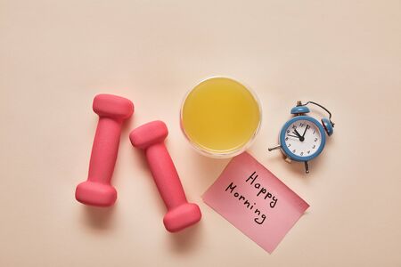 top view of sticky note with happy morning lettering, pink dumbbells, orange juice and small alarm clock on beige backgroundの写真素材