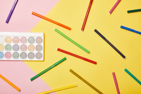 top view of multicolored felt pens and watercolor paints on pink and yellow background