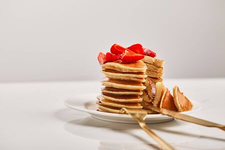 selective focus of pancakes with strawberries and golden cutlery on white plate isolated on greyの写真素材