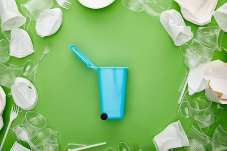 top view of blue recycle bin between crumpled plastic cups, forks, plates and cardboard container on greenの写真素材