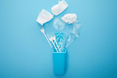 top view of crumpled plastic cups, forks and spoons near recycle bin on blue background