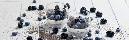 panoramic shot of tasty yogurt with chia seeds, blueberries, blackberries near scattered seeds and berries on white wooden isolated on blackの写真素材