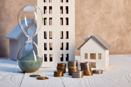 houses models on white wooden table near hourglass and coins, real estate concept
