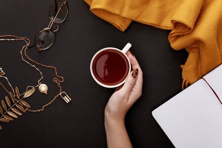 partial view of female hand near tea in mug and autumnal accessories on black background