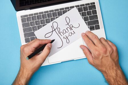 cropped view of man writing on card with thank you lettering near laptop on blue backgroundの写真素材