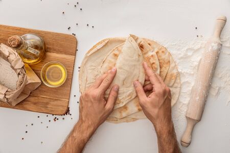 cropped view of man wrapping flat lavash bread near rolling pin and wooden cutting board with flour...