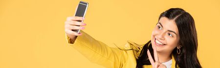 happy girl showing peace sign while taking selfie on smartphone, isolated on yellow