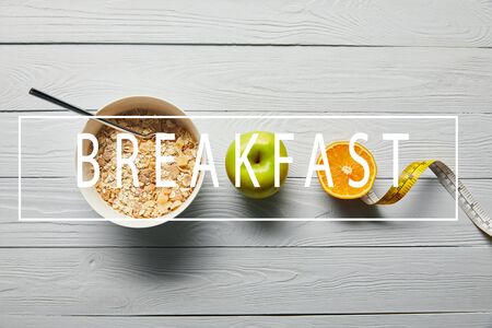 flat lay with breakfast cereal in bowl, apple, orange and measuring tape on wooden white background with breakfast letteringの写真素材
