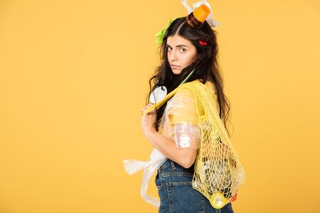 confused girl with trash in hair holding bag with rubbish isolated on yellow