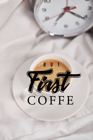top view of coffee in white cup on saucer near silver alarm clock on bedding with but first coffee...