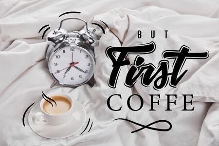 coffee in white cup on saucer near silver alarm clock in bed with but first coffee illustrationの写真素材
