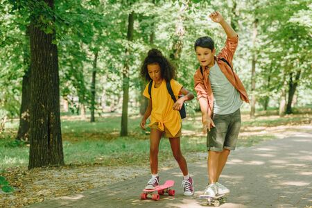 two multicultural schoolkids riding skateboards in green sunny parkの写真素材