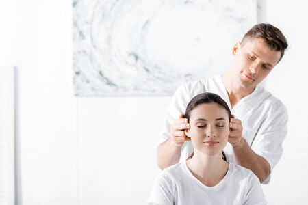 young woman sitting with closed eyes while masseur touching her earsの写真素材
