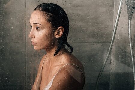 beautiful sad woman in shower at home through glass with water drops