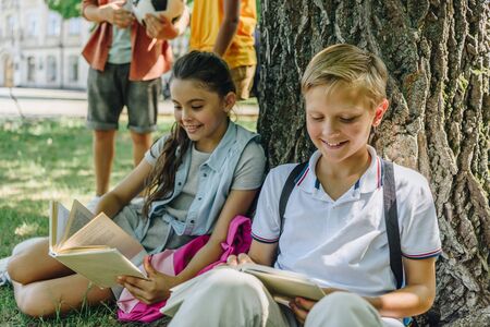 adorable schoolchildren sitting on lawn and reading books near multicultural friendsの写真素材
