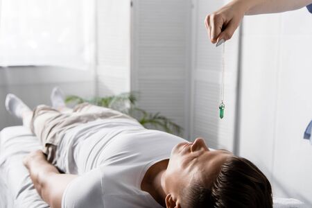 cropped view of hypnotist standing near man on massage table and holding green stone