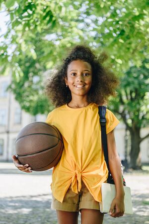 cheerful african american schoolgirl holding ball and smiling at cameraの写真素材