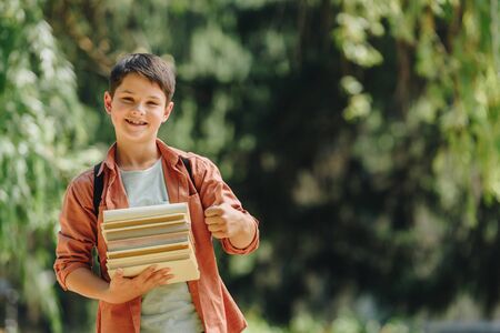 cheerful schoolboy smiling at camera while showing thumb up and holding booksの写真素材