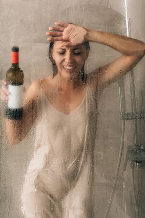 lonely depressed woman holding wine bottle, touching glass door and crying in showerの写真素材