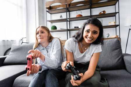 KYIV, UKRAINE - JULY 10, 2019: multicultural friends sitting on sofa and playing video game in...