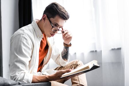 handsome man in shirt and glasses reading book in apartmentの写真素材