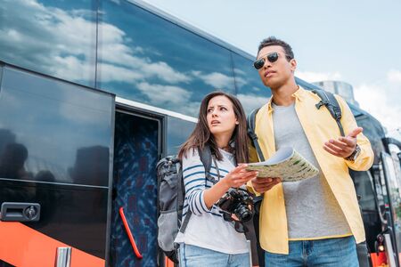 Woman with digital camera near mixed race friend with map and backpack