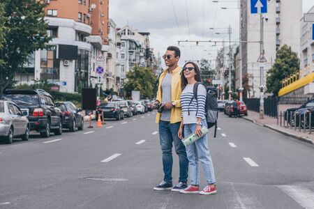 Woman holding map and standing on road near bi-racial friend with backpack