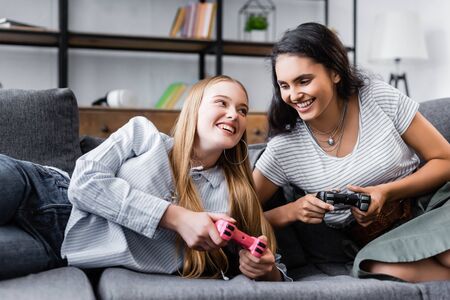 KYIV, UKRAINE - JULY 10, 2019: multicultural friends sitting on sofa and playing video game in...