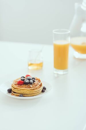 selective focus of plate with tasty pancakes and berries near glass and jug of orange juice and jar with honey on white tableの写真素材