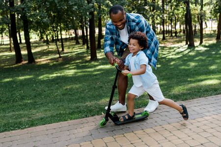 cheerful african american father near happy kid on scooterの写真素材