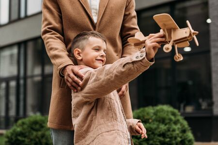 partial view of dad in coat and smiling boy playing with wooden toy plane on street