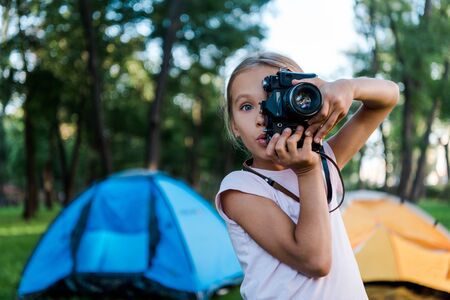 surprised kid holding digital camera while taking photo near campsの写真素材