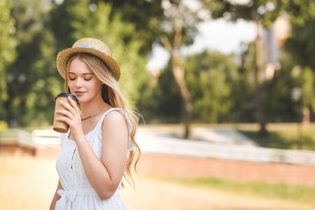 beautiful girl in white dress and straw hat holding paper cup and drinking coffee with closed eyesの写真素材