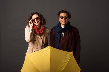 happy stylish interracial couple in autumn outfit holding yellow umbrella on black background