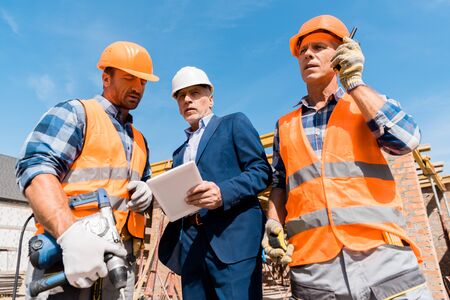 low angle view of builder holding walkie talkie near coworker and businessman with digital tablet