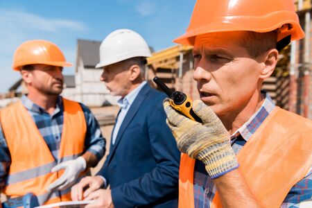 selective focus of builder holding walkie talkie while talking near coworker and businessman