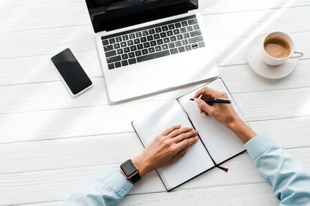 top view of woman holding pen while writing in notebook near laptop and cup of coffee