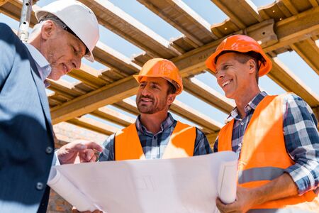 low angle view of builders near mature businessman in helmet with blueprint