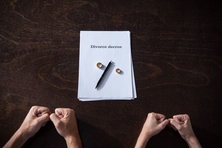 partial view of couple clenching fists at table with divorce documents and rings