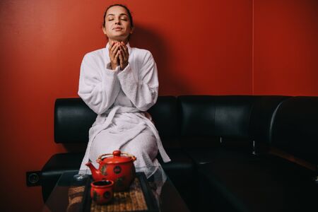 attractive woman in white bathrobe with closed eyes sitting on sofa and holding cupの写真素材
