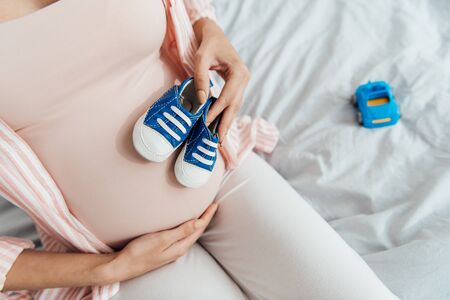 partial view of pregnant woman with toy car and baby shoes sitting on bed and touching belly