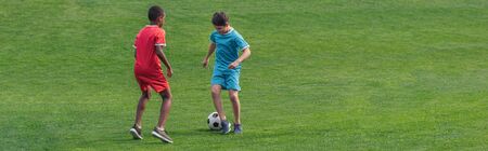 panoramic shot of cute multicultural kids playing football on grass